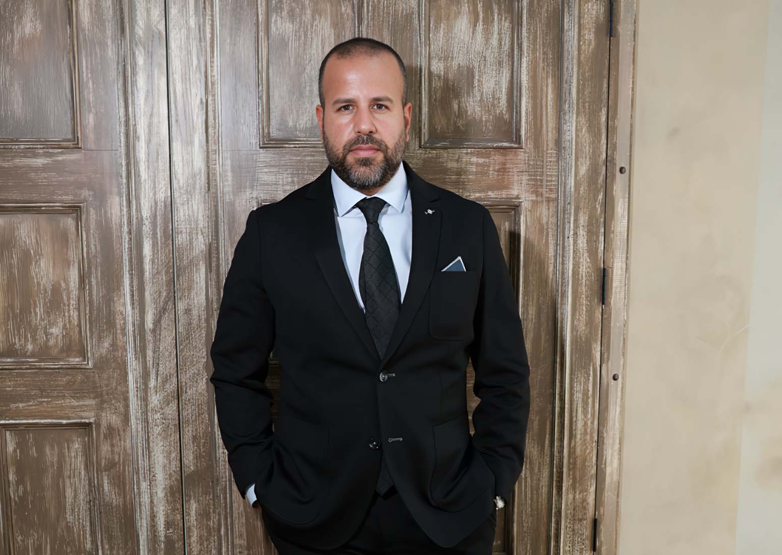 Man in a tailored black suit and tie stands with hands in pockets in front of weathered wooden doors.