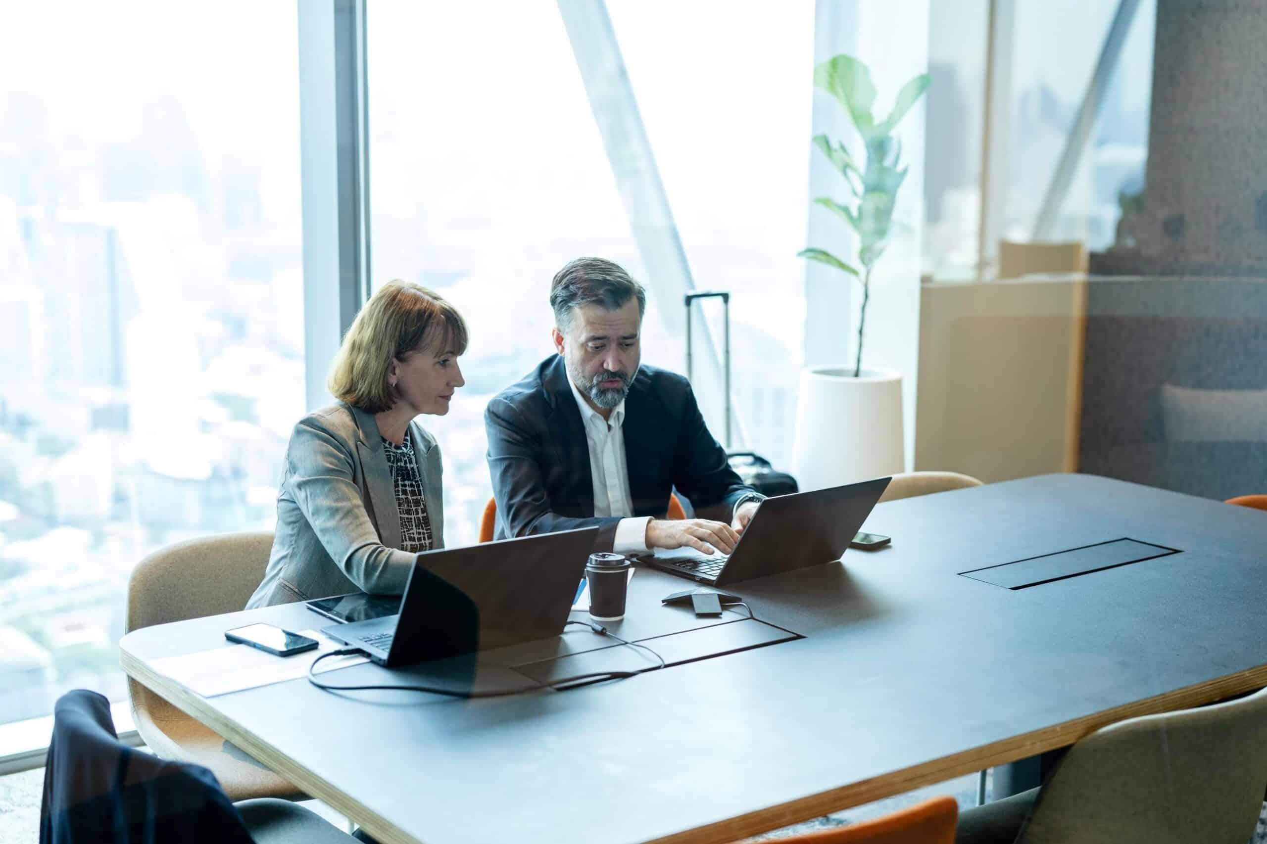 Two professionals in business attire collaborate over laptops at a conference table in a bright office.